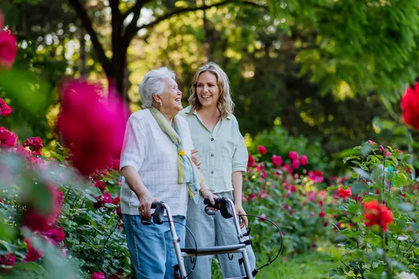 Eine alte Frau mit Rollator und eine junge Frau sind zusammen in einem von roten Rosen übersäten Park. Die junge Frau stützt den Arm der alten Frau. Beide lachen und sehen glücklich aus.