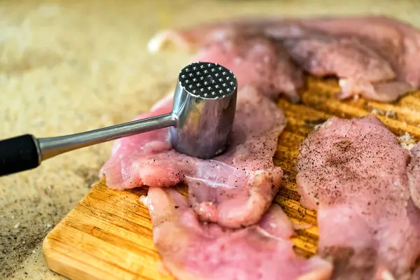 Rohe Schnitzel, mit Fleischzartmacher gewürzt, liegen auf einem Brett. Ein Fleischklopfer ruht auf einem Schnitzel.