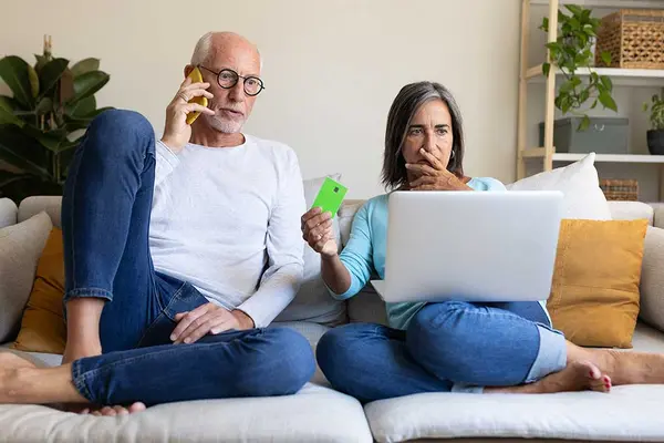 Ein Mann und eine Frau sitzen auf dem Sofa, der Mann telefoniert, die Frau hält erschrocken eine Kreditkarte hoch 