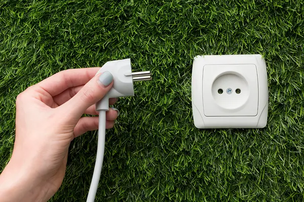 Eine Hand hält einen weißen Netzstecker neben eine Steckdose, die auf einer Fläche aus grünem Kunstrasen angebracht ist.
