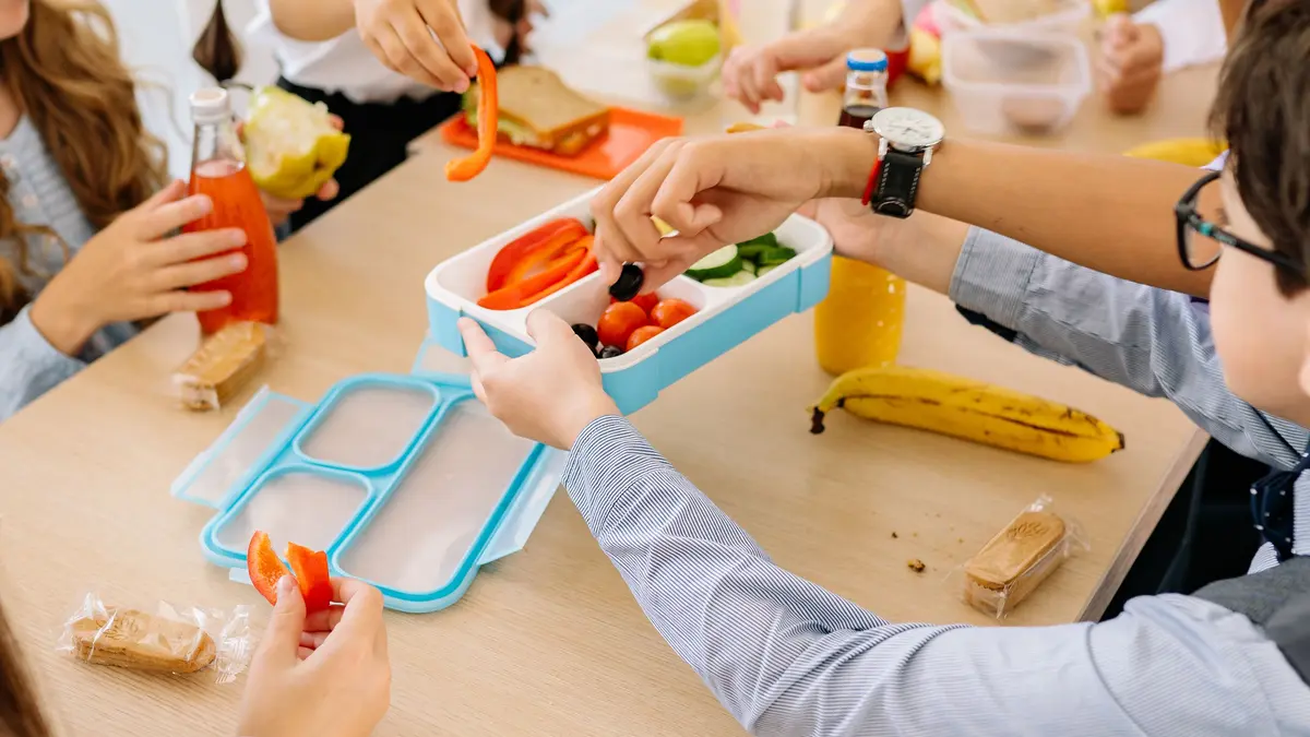 An einem Tisch sitzen 4 Kinder. Alle greifen nach einer hellblauen Brotbox, in der sich verschiedenes Gemüse befindet. Auf dem Tisch sind Lebensmittel, wie belegte Brote sowie Obst und Gemüse verteilt. In Glasflaschen mit verschiedenen Säften und Wasser stehen auch auf dem Tisch.