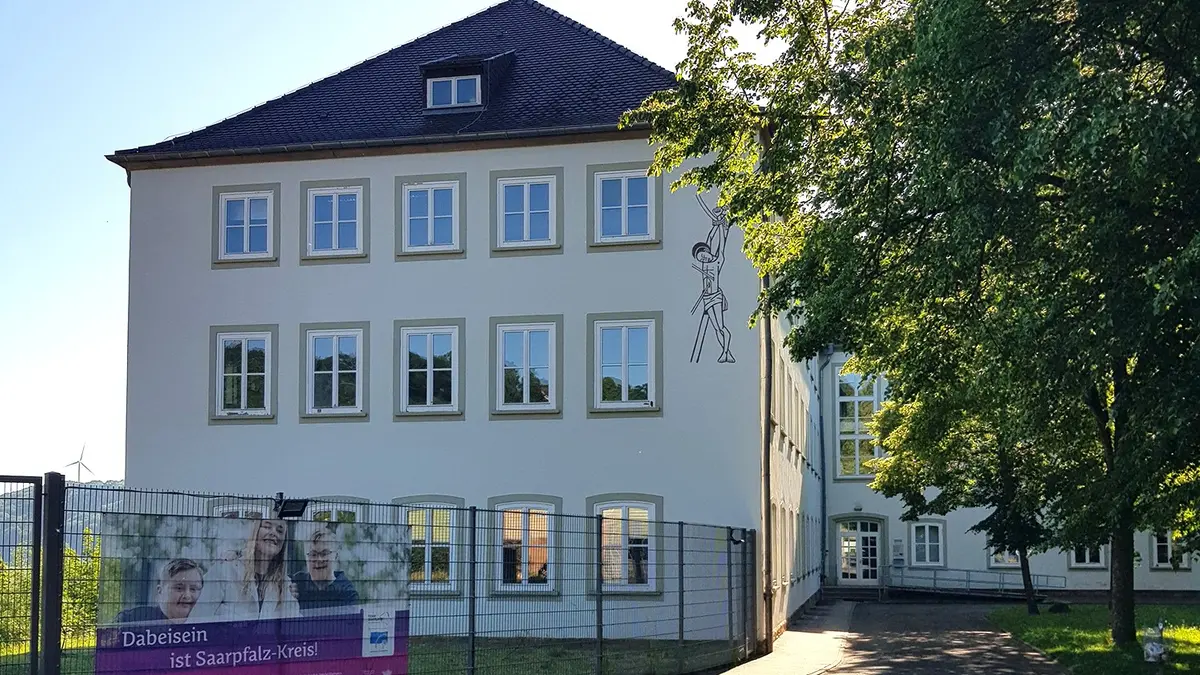 Stirnseite eine dreistöckigen beigen Gebäudes mit Dach. Je Stockwerk fünf Kassettenfenster. Mannshoher Stabzaun mit Ankündigungstransparent. Rechts davon Innenhof mit großem Baum, durch den sich Sonnenlicht bricht.