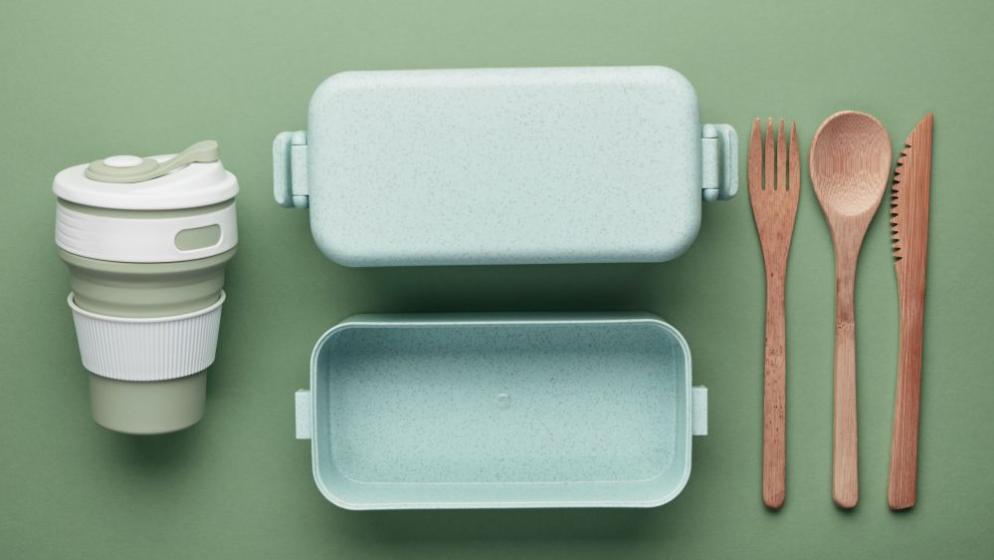 Foto von einem To-Go-Becher, einem Brotdose und Besteck aus Bioplastik und Holz, von oben fotografiert auf grünem Untergrund.