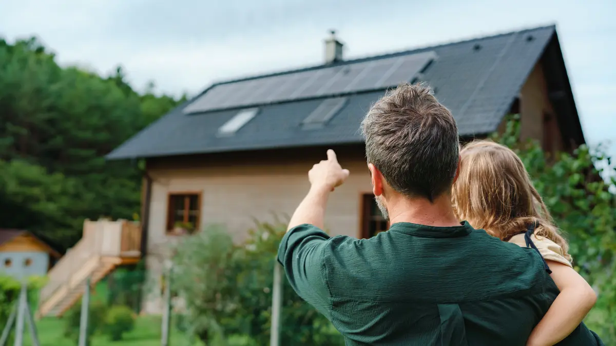 Vater mit Kind auf dem Arm zeigt auf Haus mit Photovoltaik Dach