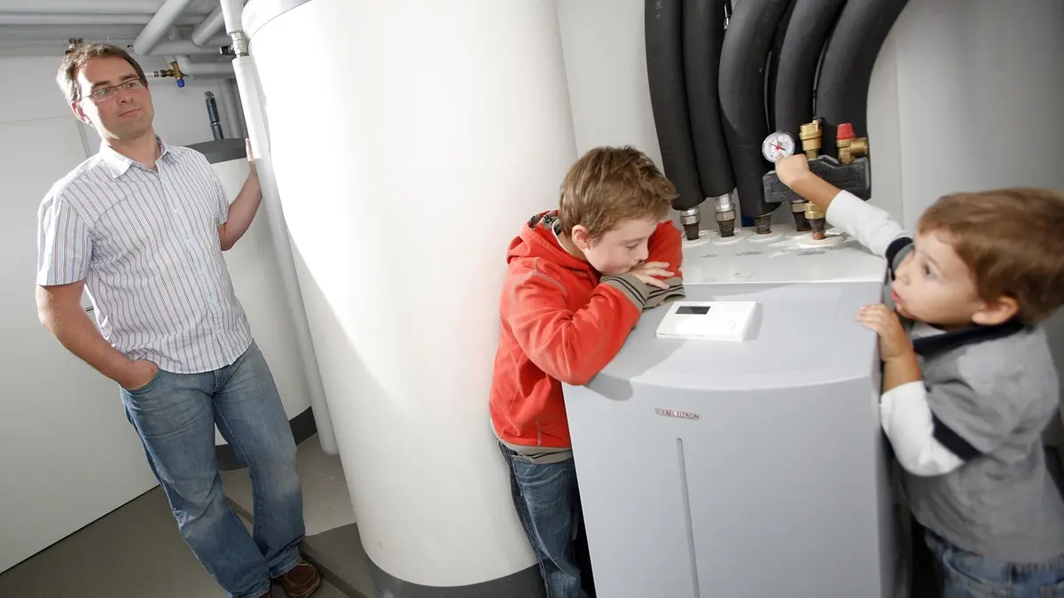 Ein Vater steht im Keller seines Hauses neben seiner Wärmepumpe. Zwei Kinder sind auch anwesend.