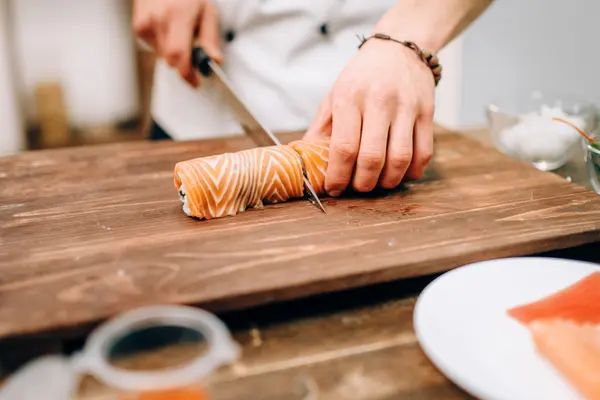Eine mit Lachsscheiben ummantelte Sushirolle liegt auf einem Holzbrett. Sie wird von einer Hand gehalten, die andere Hand hält ein großes Messer, mit dem sie die Rolle in der Mitte durchschneidet.