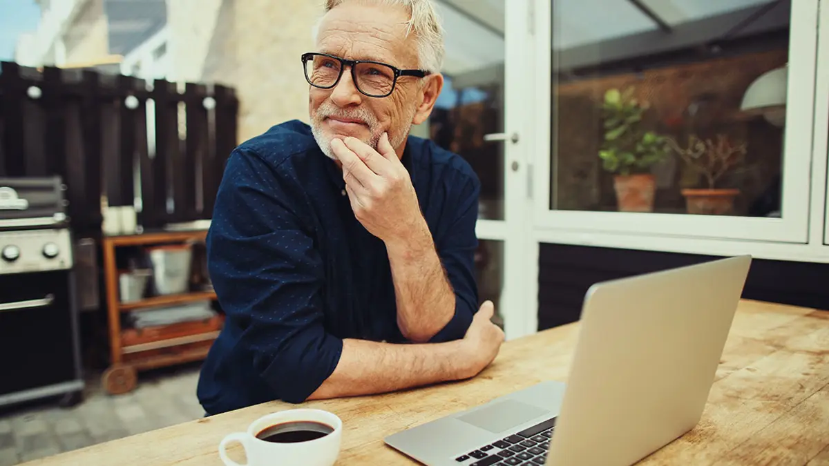 Im älterer Mann sitzt nachdenklich auf der Terrasse eines Hauses, vor ihm ein Laptop und eine Tasse Kaffee