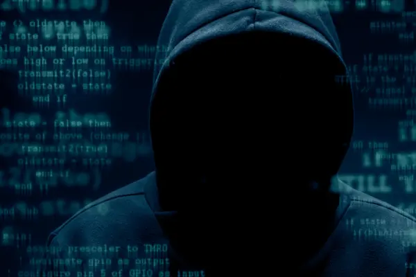 eine Person in dunklem Kapuzenpullover, dessen Gesicht im Dunkeln ist, steht vor eine Wand, auf der Codes laufen
