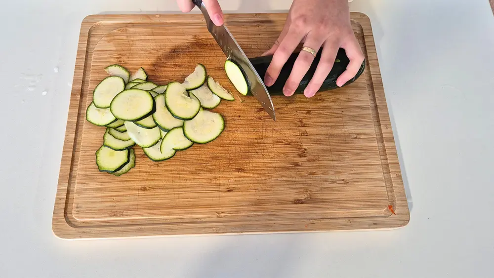 Eine Person schneidet eine Zucchini auf einem Holzbrett in dünne Scheiben. Die bereits geschnittenen Scheiben liegen bereit, während die Hand mit einem Küchenmesser weitere Scheiben schneidet. Im Hintergrund sind ein weißer Tisch und ein Holzfußboden zu sehen.