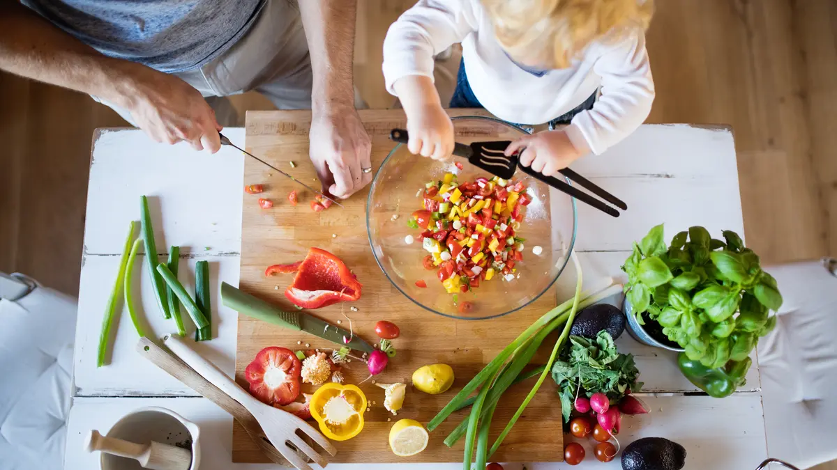 Aus der Vogelperspektive bereiten ein Erwachsener und ein Kind gemeinsam einen bunten Salat mit frischem Gemüse und Kräutern zu. Auf dem Tisch liegen verschiedene Zutaten wie Paprika, Tomaten, Radieschen, Frühlingszwiebeln, Basilikum und Avocado, während in einer Schüssel bereits gemischtes Gemüse liegt.