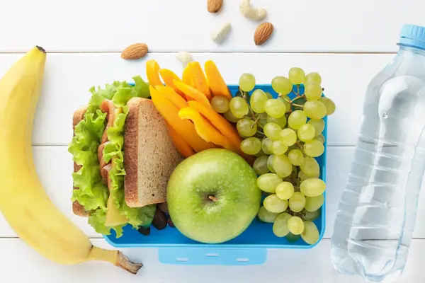 In einer Brotzeitbox liegen ein belegtes Brot, ein grüner Apfel, Paprika-Sticks und grüne Weintrauben. Links daneben liegt eine Banane. Rechts neben der Box liegt eine Flasche mit Wasser.