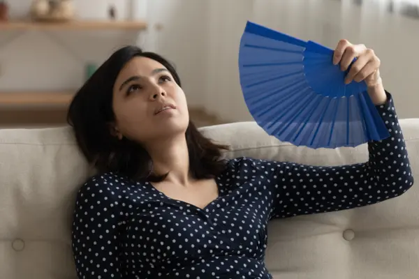 Eine Frau sitzt vor ihrem Sofa und fächert sich mit einem Fächer Luft ins Gesicht.