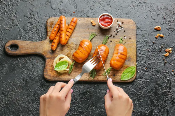 ein Holzbrett mit vegetarischen Würstchen, Ketchup und etwas Gemüse, zwei Hände halten Besteck