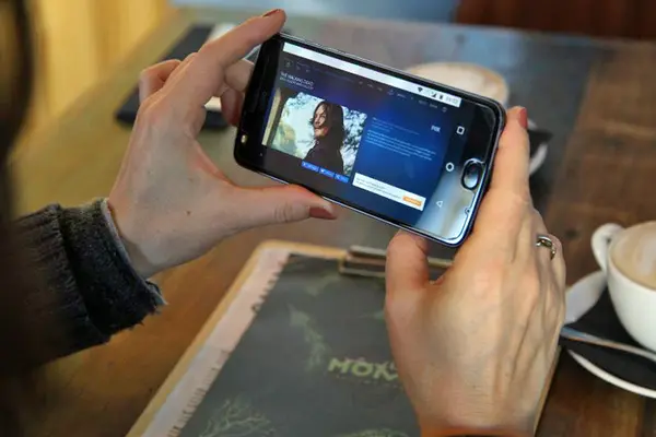 Eine Frau hält in einem Café ein Smartphone in der Hand, auf dem ein Video-Streaming-Dienst zu sehen ist.