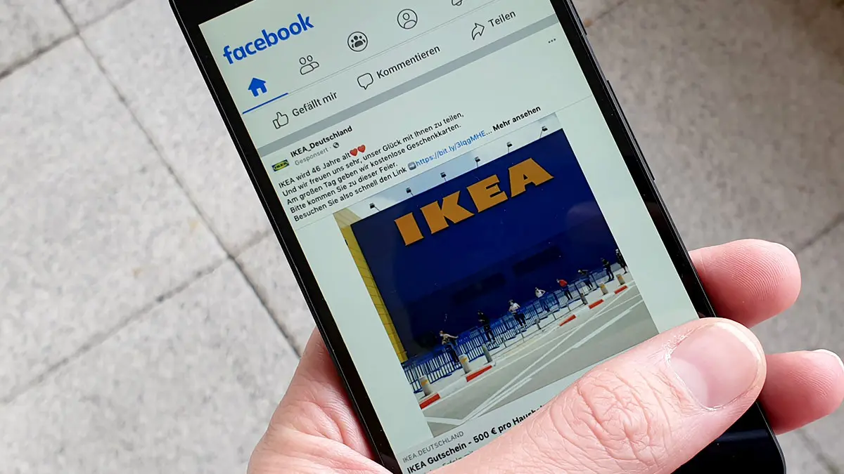 Hand hält Smartphone, Display zeigt einen Beitrag mit dem Foto eines Ikea-Gebäudes auf Facebook