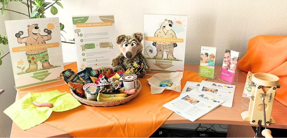 Tisch mit Flockenquetsche, Banane, Möhre, Chipstüte und weiteren Materialien wie Flyer und 