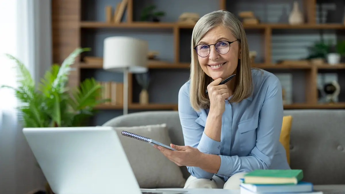 Eine Frau mittleren Alters mit Brille sitzt auf dem Sofa, vor ihr steht ihr Laptop und ein Stapel Bücher. Sie hält ein Notizblock in der einen Hand und einen Kugelschreiber in der anderen. Diese Hand hat sie an ihr Kinn gestützt und lacht freundlich in die Kamera.