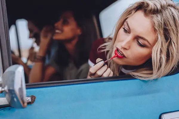 Zwei Frauen in einem blauen Auto, eine malt sich die Lippen an