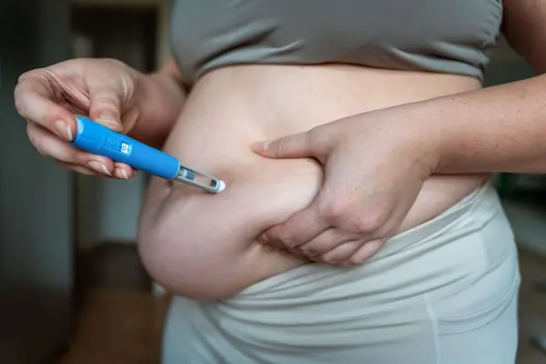 Eine Nahaufnahme zeigt eine Person, die sich mit einem blauen Injektionspen (ähnlich einem Insulin- oder Abnehmspritzen-Stift) ein Medikament in das Bauchfett injiziert. Die Person trägt Sportkleidung und drückt mit einer Hand eine Hautfalte am Bauch zusammen, während sie mit der anderen Hand den Pen senkrecht ansetzt. Der Hintergrund ist leicht unscharf und zeigt einen Innenraum.