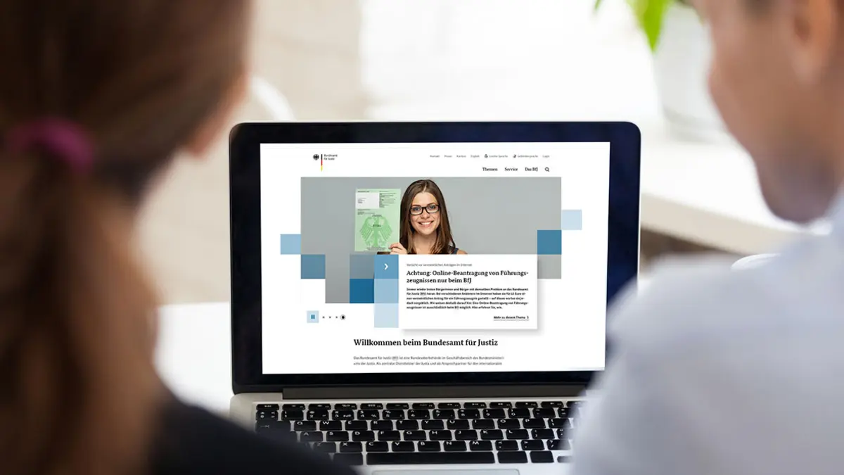 Ein Mann und eine Frau sitzen vor einem Laptop, auf dem Bildschirm die Seite des Bundesjustizamts