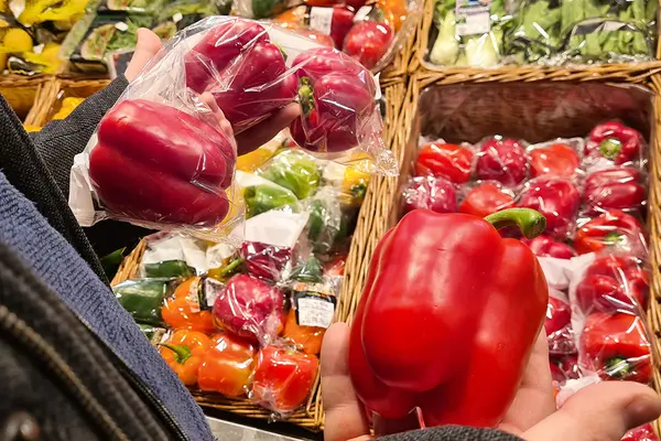 Im Supermarkt hält jemand unverpackte und in Plastikfolie verpackte rote Paprika in der Hand