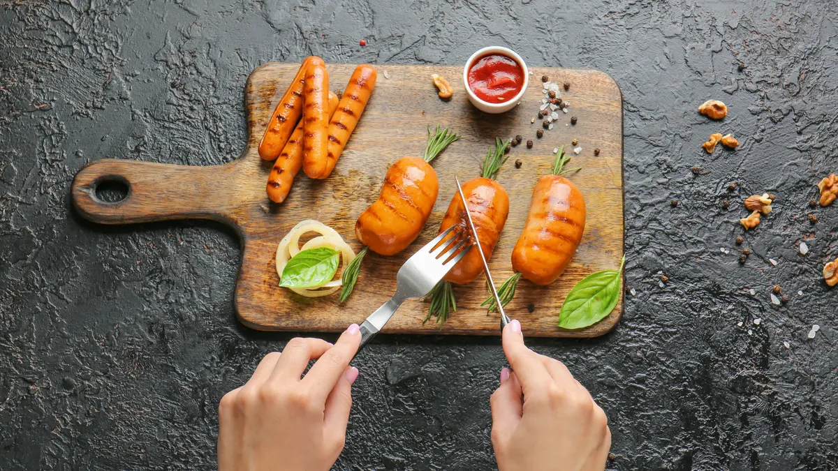 ein Holzbrett mit vegetarischen Würstchen, Ketchup und etwas Gemüse, zwei Hände halten Besteck