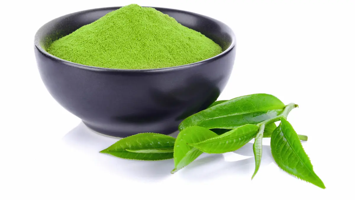 Grünes Matcha-Pulver in einer schwarzen Keramikschale und Teeblätter davor.