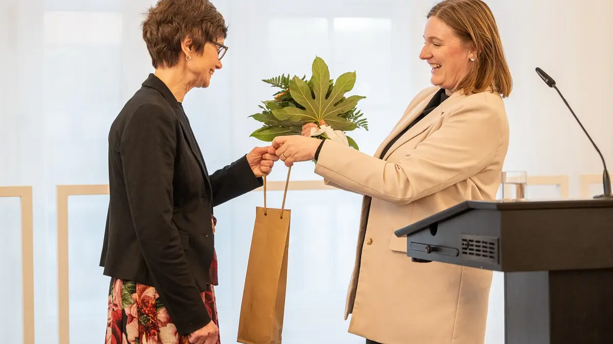 Rechts steht eine Frau an einem Rednerpult und überreicht einer anderen Frau links im Bild einen Blumenstrauß und eine Geschenktüte.