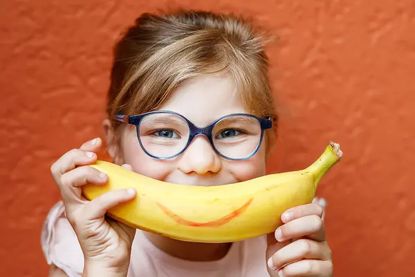 Ein Kind hält sich eine Banane mit aufgemaltem Lächeln vor den Mund