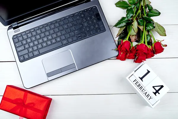 Neben einem Laptop liegen roten Rosen und ein verpacktes Geschenk auf einem Tisch.
