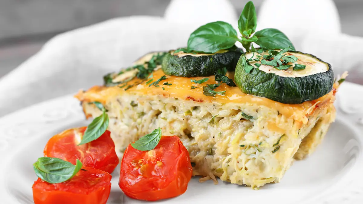 Ein Stück Gemüsetarte auf einem Teller, garniert mit Zucchini, Tomate und Basilikum