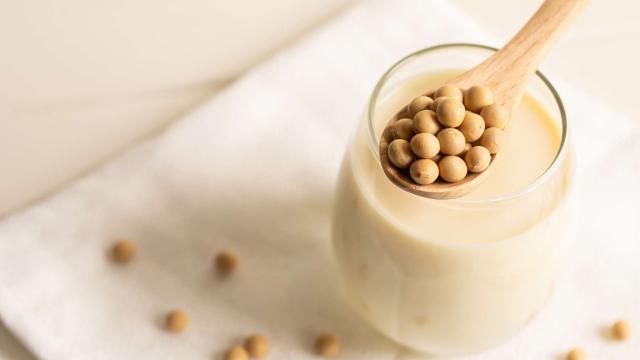 Ein Glas cremige Sojamilch steht im Zentrum dieser hellen, minimalistischen Komposition auf einem weißen Stoffuntergrund, während ein darüber balancierter Holzlöffel eine Häufung runder, gelblicher Sojabohnen präsentiert. Die sanfte Beleuchtung in warmen Beigetönen betont die natürliche Textur der Zutaten, die durch weitere dekorativ verstreute Bohnen im weichgezeichneten Hintergrund ergänzt werden.