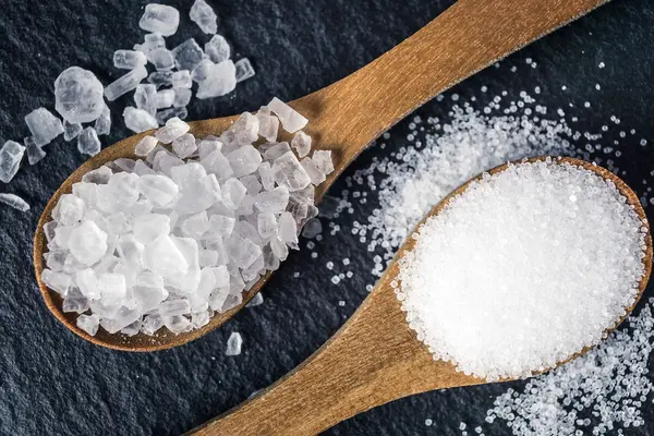 Zwei Holzlöffel je mit grobem und feinem Salz liegen auf einer schwarzer Granitplatte, umgeben von verstreuten Salzkörnern.