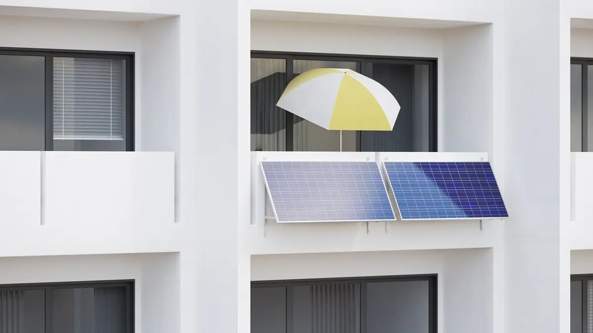 Ein Mehrfamilienhaus. An einem Balkon hängt ein Balkonkraftwerk und steht ein Sonnenschirm.