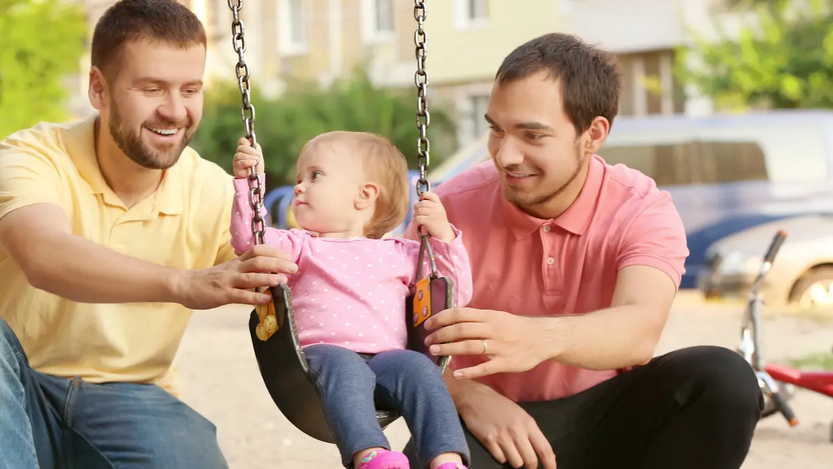 Zwei Männer spielen mit ihrer Tochter, die auf einer Schaukel sitzt.