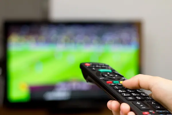 Foto von einer Hand, die eine Fernbedienung hält und auf einen Fernsehbildschirm richtet, auf dem ein Fußballspiel läuft