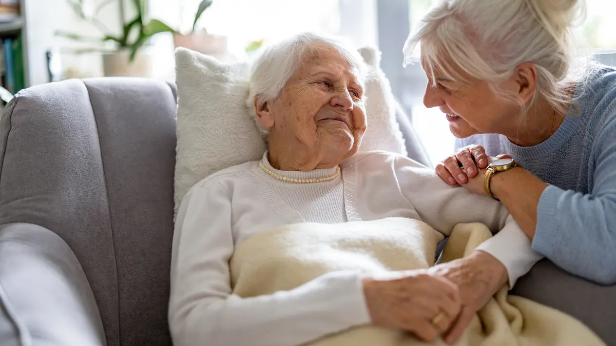 Eine Seniorin Sitzt durch ein Kissen gestützt auf der Couch und lächelt. Eine weitere Frau lehnt sich liebevoll zu ihr rüber und schaut sie an.