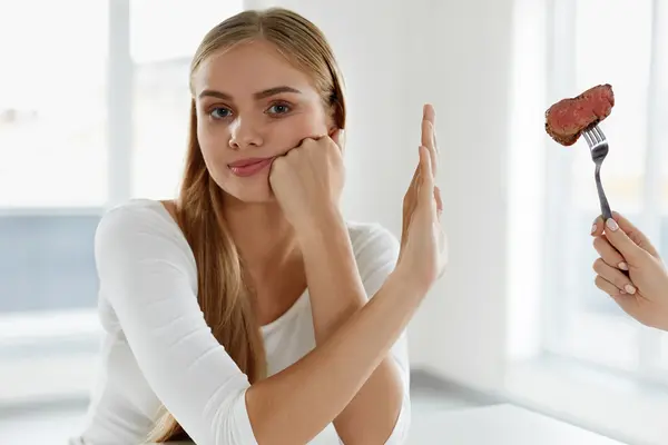 Frau hält Hand ablehnend vor Gabel, die ihr mit einem Stück Fleisch von der Seite gereicht wird