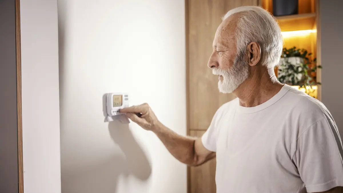 Ein älterer Mann bedient eine Klimaanlage/ Heizung an der Wand in seinem Wohnraum.