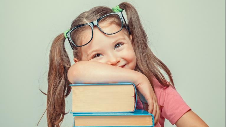 Mädchen mit Brille hat den Arm über einen Stapel Bücher gelegt.