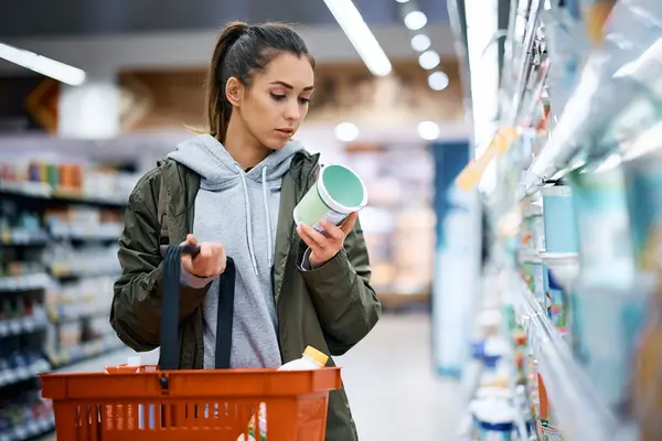 Eine Frau steht im Supermarkt und schaut kritisch auf eine Verpackung eines Lebensmittel. 