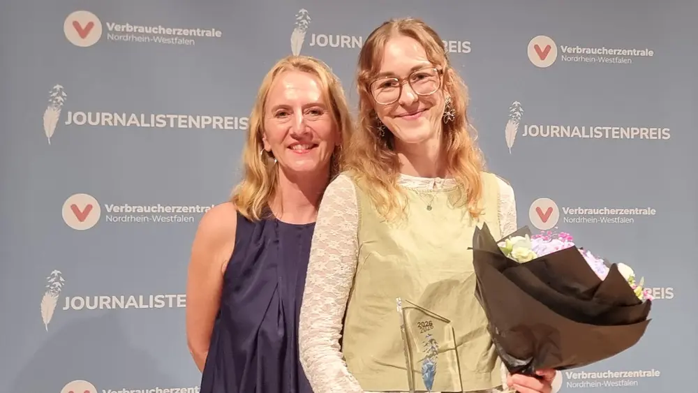 Zwei Frauen stehen nebeneinander und lächeln in die Kamera. Eine von ihnen hält einen Blumenstrauß und eine Trophäe in der Hand.