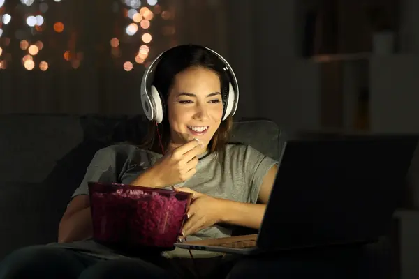 Eine lächelnde Frau mit weißen Kopfhörern und einer Schale Popcorn , die nachts im Internet Fernsehen schaut.