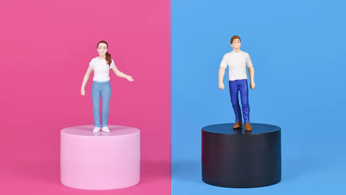 Zwei Spielfiguren, eine weibliche und eine männliche, stehen auf getrennten Sockeln vor einem geteilten Hintergrund. Die Frau steht auf einem rosa Sockel vor einem rosa Hintergrund, während der Mann auf einem schwarzen Sockel vor einem blauen Hintergrund steht. Beide tragen weisse T-Shirts und blaue Hosen.