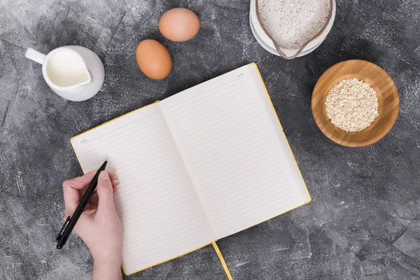 Hand mit Stift und einem leeren Notizbuch, Eier, Milch, Haferflocken an der Seite um das Notizbuch herum.