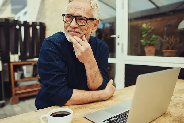 Im älterer Mann sitzt nachdenklich auf der Terrasse eines Hauses, vor ihm ein Laptop und eine Tasse Kaffee