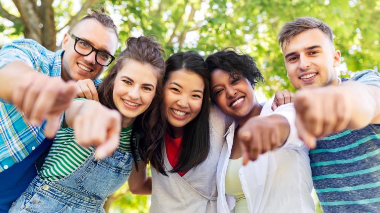 Grupp aus 5 jungen Menschen schaut in die Kamera und zeigt mit Fingern auf Kamera.