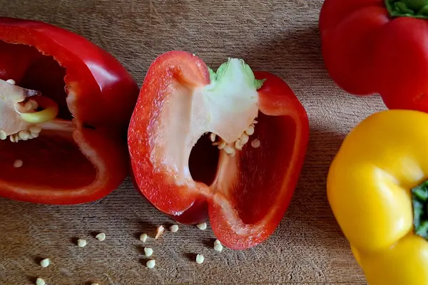 Eine rote Paprika liegt halbiert auf dem Tisch, vor ihr liegen Paprikakerne. Daneben befinden sich je eine ganze rote und eine ganze gelbe Paprika.