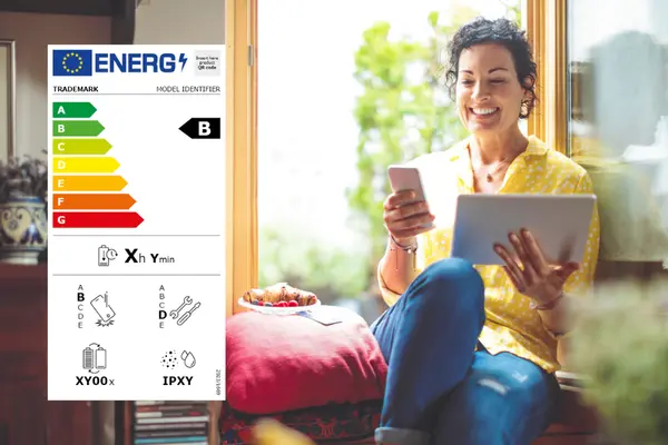 Frau am Fenster mit Tablet und Smartphone in der Hand, links im Bild das Energielabel für Smartphones und Tablets