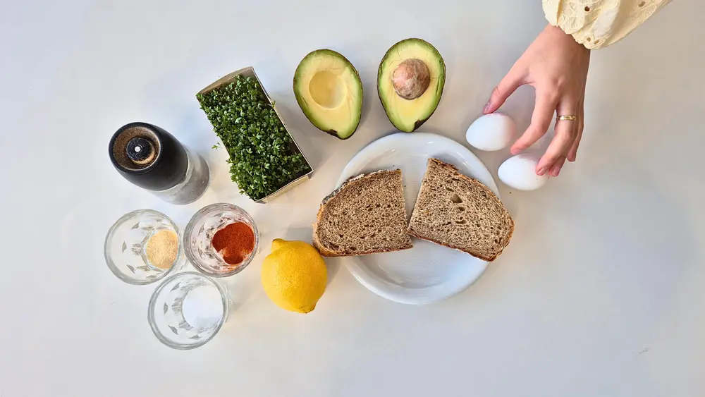 Zutaten auf einem weißen Tisch: zwei Eier, eine Zitrone, Salz, Pfeffer, Paprikapulver, Knoblauchpulver, eine halbierte Avocado, frische Kresse und zwei Scheiben Vollkornbrot.